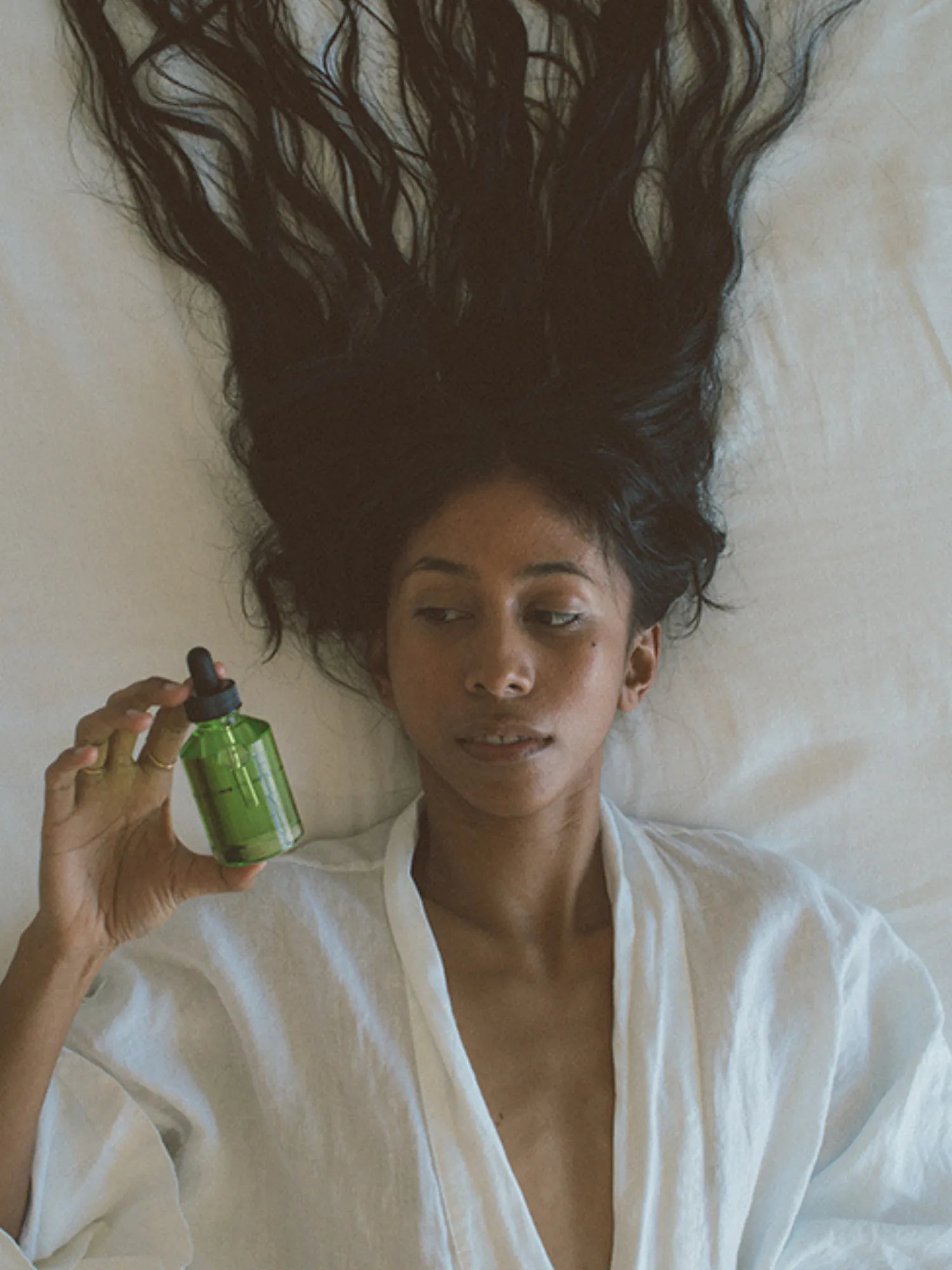 Woman lying on a bed holding HINU Hair Growth Oil beside her long hair