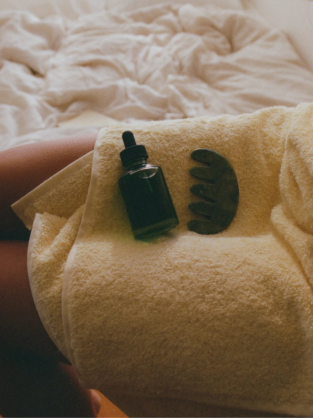 HINU Hair Growth Oil and jade tool resting on a woman's towel-covered lap
