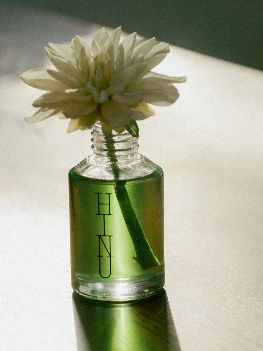 A clear glass bottle filled with translucent green liquid, featuring the vertical black text 'HNU' on the front, holding a single white flower. The bottle is placed on a reflective light-colored surface with a soft shadow.