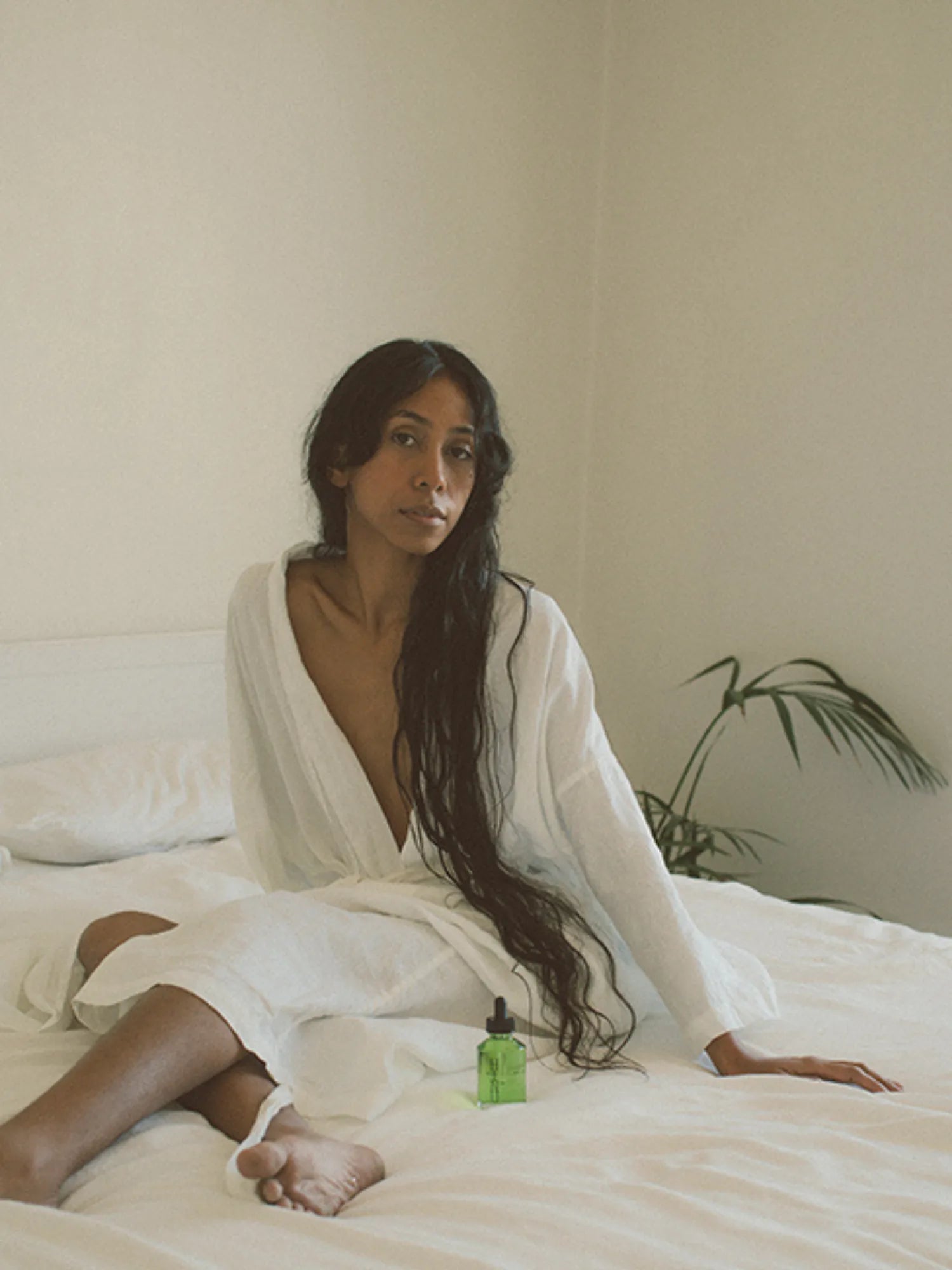 Woman in a white robe seated on a bed beside HINU Hair Growth Oil
